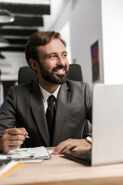 Foto de hombre de negocios feliz en traje formal trabajando con documentos y portátil mientras está sentado en el escritorio en la oficina de planta abierta