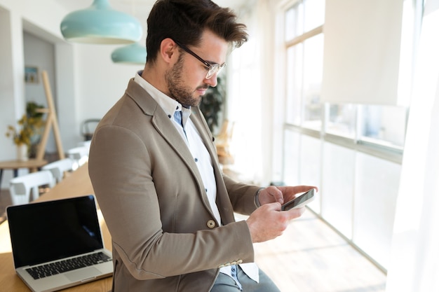 Foto de hombre de negocios joven y guapo con su teléfono inteligente mientras está sentado en la mesa en la oficina.