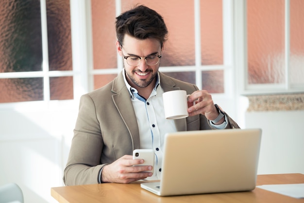 Foto de hombre de negocios joven guapo con su teléfono inteligente mientras toma café frente a la computadora portátil en la oficina.