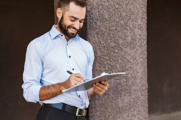 Foto de un hombre de negocios joven sonriente alegre feliz de pie cerca del centro de negocios sosteniendo notas de escritura del portapapeles.