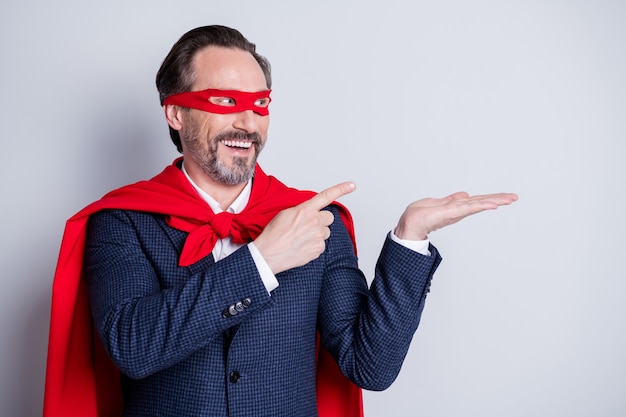 Foto de hombre de negocios maduro, alegre y confiado, disfraz de superhéroe, dedos directos, brazo abierto, retención de producto novedoso, espacio vacío, oferta, traje, máscara facial roja, manto aislado fondo gris