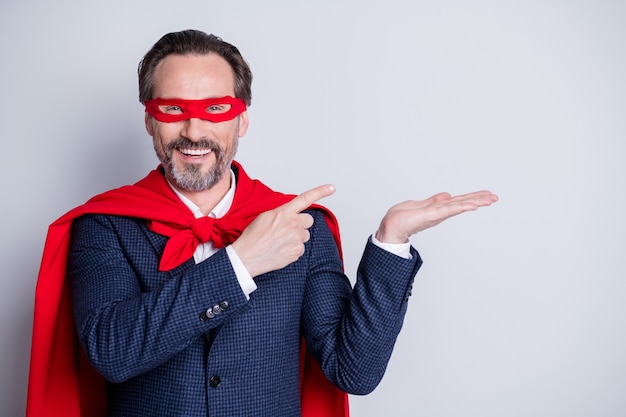 Foto de hombre de negocios maduro de confianza, disfraz de superhéroe, dedos directos, brazo abierto, retención de producto novedoso, espacio vacío, oferta, traje de vestir, máscara facial roja, manto aislado fondo gris