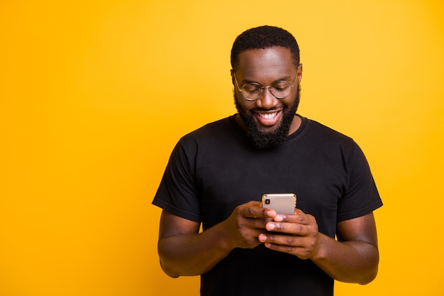 Foto de hombre negro radiante con dientes alegre navegando a través de su teléfono en busca de nueva información en espectáculos aislados pared de colores vivos