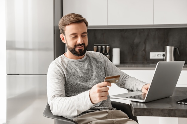 Foto de un hombre sonriente de 30 años en ropa casual sentado solo en el apartamento y usando una computadora portátil con tarjeta de crédito para comprar en línea