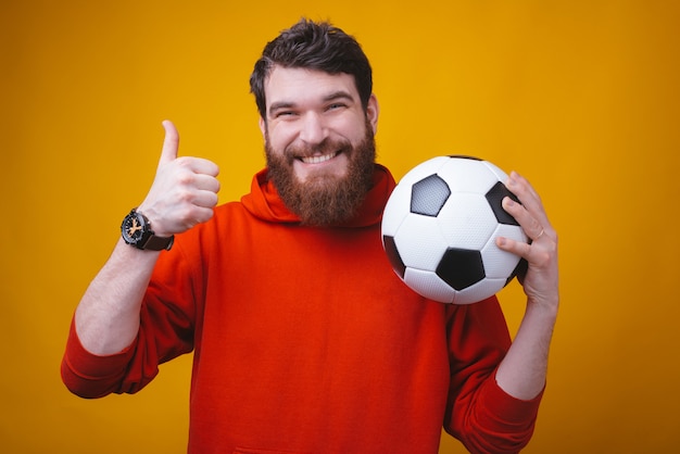 La foto del hombre sonriente está sosteniendo una pelota de fútbol o fútbol y muestra el pulgar hacia arriba o como gesto.