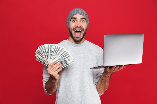 Foto de hombre sorprendido de 30 años en ropa casual con dinero en efectivo y portátil plateado aislado