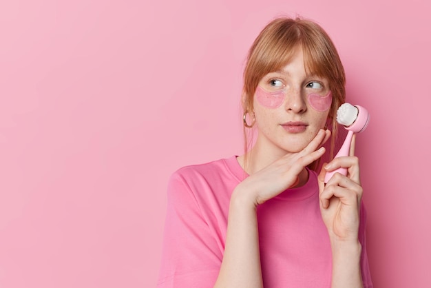 La foto horizontal de una chica pecosa pelirroja pensativa toca la línea de la mandíbula se somete suavemente a tratamientos faciales aplica parches de hidrogel debajo de los ojos sostiene el cepillo facial mira hacia otro lado aislado sobre la pared rosa del estudio