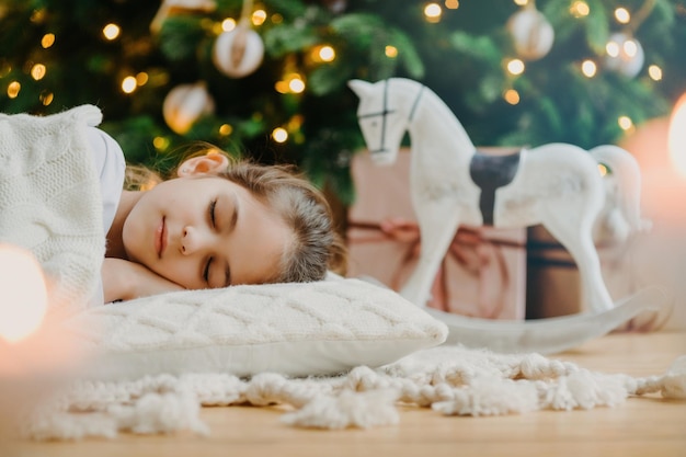 La foto horizontal de una chica relajada cubierta con un suéter blanco tiene sueños agradables yace debajo de un árbol de Navidad decorado, un balancín de juguete y cajas de regalo alrededor de Kid at home Vacaciones acogedoras Atmósfera doméstica