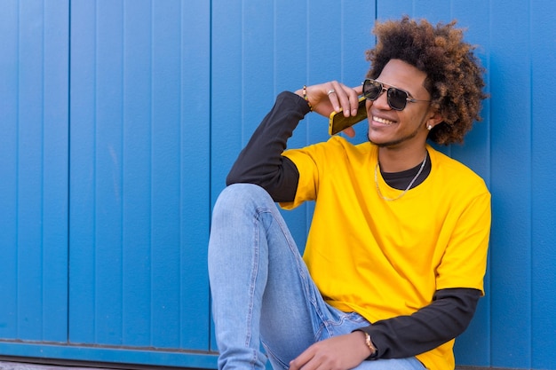 Foto horizontal com fundo azul de um homem sentado na rua conversando ao celular relaxado