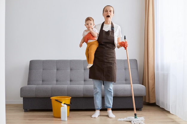 Foto horizontal da garotinha nas mãos da jovem mãe, mulher desesperada, limpando em casa, mãe exausta, cansada, limpando o chão, arrumando juntos na sala de estar