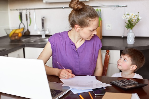 Foto horizontal da mãe trabalhadora ocupada com a documentação, faz relatório financeiro ou calcula o orçamento da família, rodeado de papéis e laptop, olha para o filho e pede para ser obediente