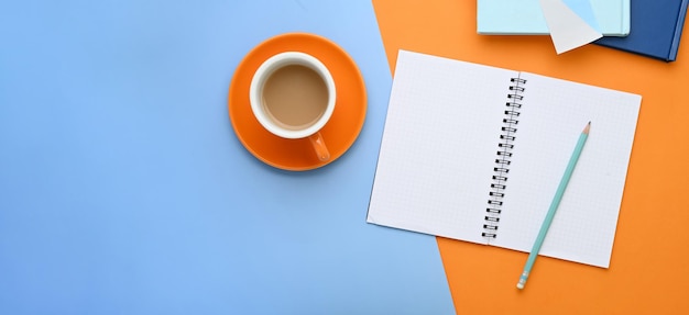 Foto horizontal de caderno, xícara de café e caneta em dois tons de fundo azul e laranja.