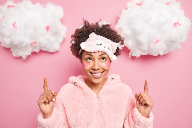 Foto horizontal de mulher alegre vestida de pijama indica acima em nuvens brancas sugere produto para dormir e descanso passa por procedimentos de beleza isolados sobre parede rosa