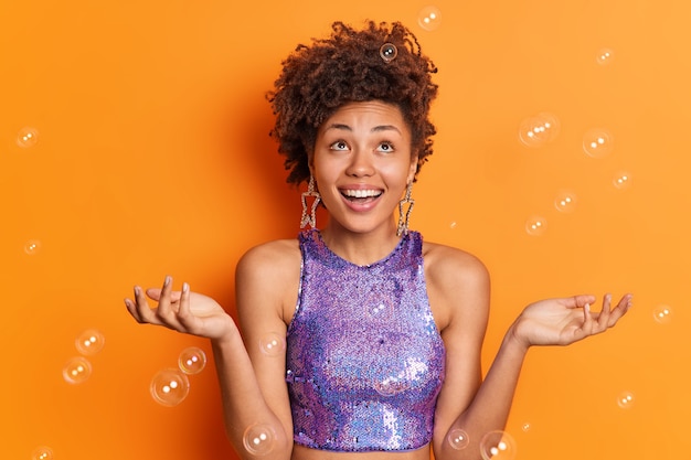 Foto foto horizontal de mulher com as palmas das mãos levantadas se sentindo muito feliz curtindo a vida usa brincos de camisa roxa da moda se prepara para a festa isolada sobre bolhas de sabão de parede laranja