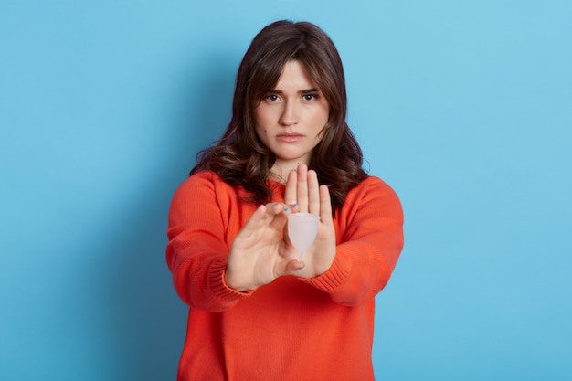 Foto horizontal de mulher morena segurando o copo menstrual de plástico na mão e mostrando o gesto de parada com a palma da mão, posando isoladas em azul