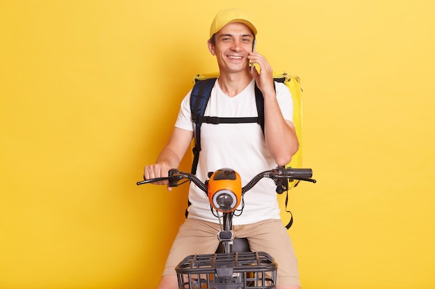 Foto horizontal de um homem de correio satisfeito e sorridente segurando pizza em caixas de papelão vestindo camiseta branca andando de scooter elétrica posando contra a parede amarela falando ao telefone com o cliente