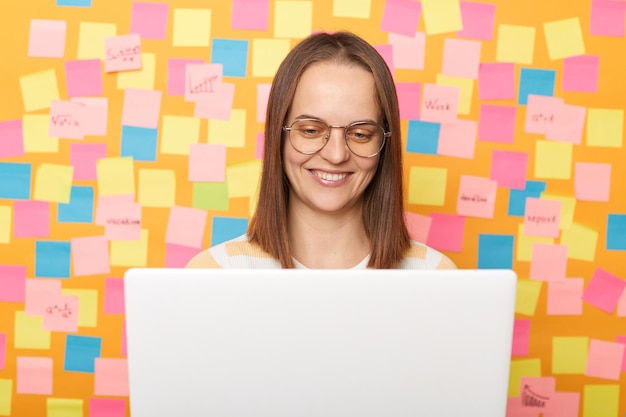 Foto horizontal de uma bela jovem adulta satisfeita vestindo camiseta posando contra a parede de papel amarelo com notas adesivas sentada na frente do laptop branco trabalhando on-line
