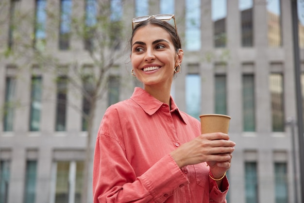 Foto horizontal de uma bela jovem sorrindo feliz curtindo o tempo de lazer caminhando ao ar livre com uma xícara de café descartável