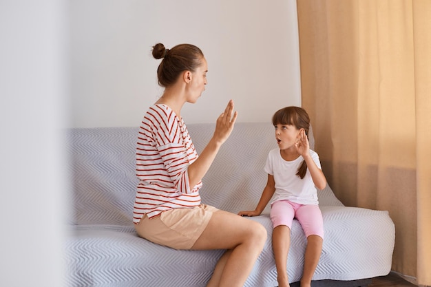 Foto horizontal de uma fonoaudióloga dando aula com uma garotinha, pessoas sentadas em um sofá confortável perto da janela, sessão de terapia com uma garota aprendendo a pronúncia dos sons