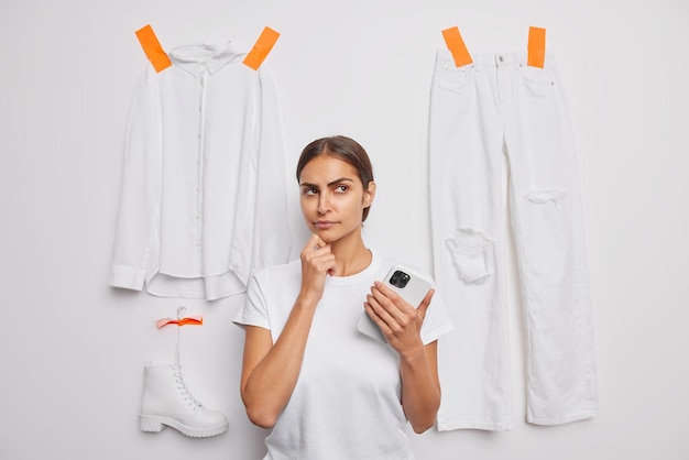 Foto horizontal de uma jovem pensativa de cabelos escuros, considerando algo com uma expressão séria segura poses de celular contra um fundo branco peças de roupa gessadas pensa no que vestir