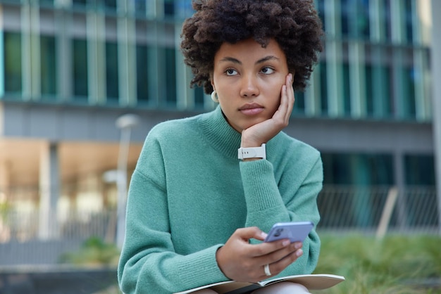 Foto horizontal de uma mulher pensativa de cabelo encaracolado aprende informações com o notebook, usa o celular para bater um papo online, usa um macacão casual e assiste a poses ao ar livre contra um fundo desfocado