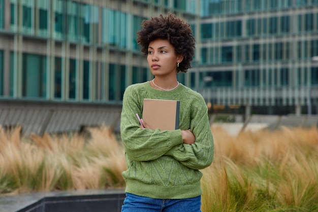 Foto horizontal de uma redatora habilidosa segura o bloco de notas e a caneta desvia o olhar e reúne pensamentos usando jeans de suéter verde poses contra um prédio de vidro moderno pondera sobre informações educacionais