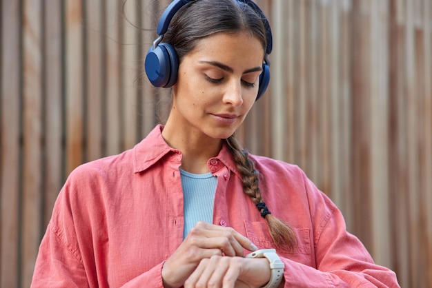 Una foto horizontal de una guapa mujer morena revisa el tiempo de guardia esperando a que alguien use audífonos estéreo en los oídos escucha una pista de audio vestida con una camisa rosa al aire libre contra un fondo borroso
