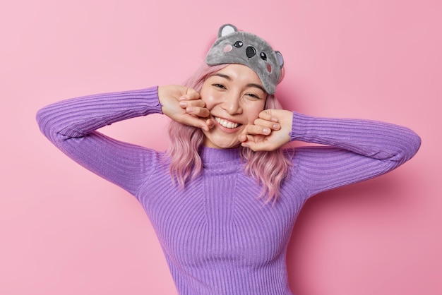 La foto horizontal de una joven asiática complacida mantiene las manos en las mejillas, sonríe suavemente, tiene una piel sana, usa una máscara para dormir y un saltador púrpura expresa emociones positivas aisladas sobre un fondo rosado.
