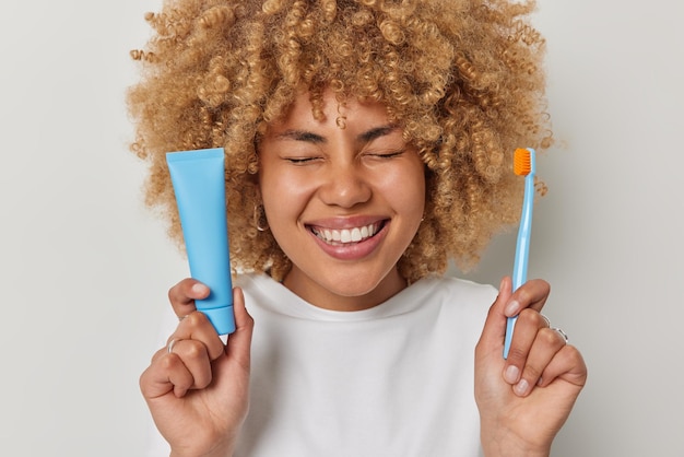 La foto horizontal de una mujer alegre con el pelo rubio y rizado sonríe con dientes y muestra dientes blancos y perfectos sostiene el cepillo de dientes y la pasta de dientes vestida con una camiseta informal posa en el interior Concepto de higiene bucal