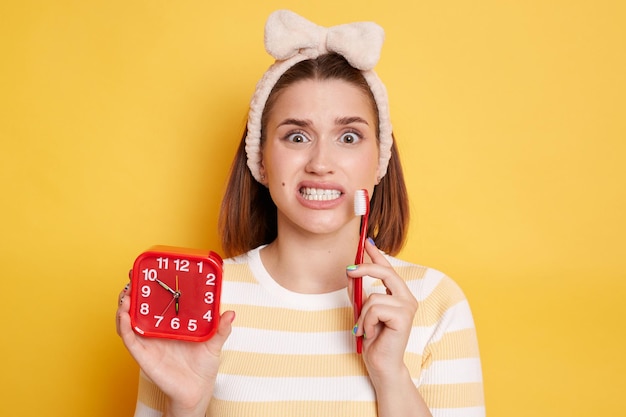 Una foto horizontal de una mujer confundida con el pelo oscuro y rizado muestra dientes blancos y perfectos, sostiene un cepillo de dientes y un reloj despertador vestido con una camisa casual que posa en el interior aislado sobre un fondo amarillo