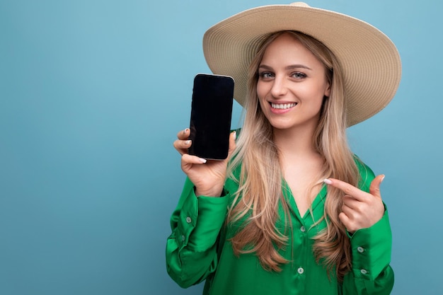 Foto horizontal de una mujer encantadora de vacaciones con un sombrero de paja de verano que muestra la pantalla del teléfono con mocup