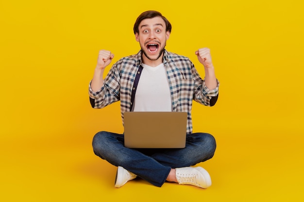 Foto in voller Größe eines glücklichen jungen Mannes, der einen Laptop hält, sitzt auf einem Boden und freut sich über den Sieg einzeln auf gelbem Hintergrund