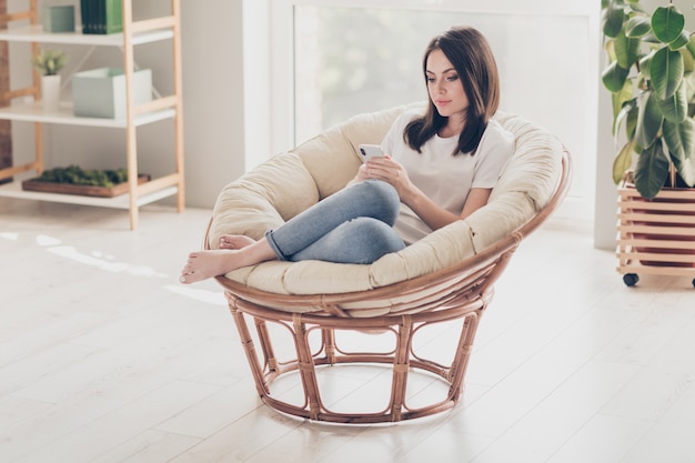 Foto in voller Größe von einem charmanten Mädchen sitzen auf einem Korbstuhl und chatten mit Freunden auf dem Smartphone im Haus im Haus