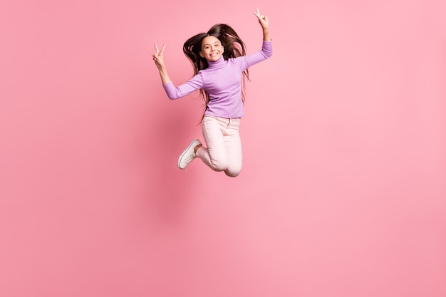 Foto in voller Größe von fröhlichen kleinen Mädchen springen lassen V-Zeichen tragen lila Pullover isoliert über pastellfarbenem Hintergrund