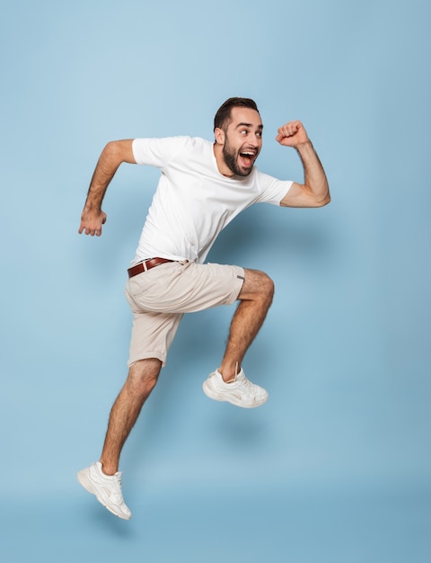 Foto in voller Länge von einem fröhlichen kaukasischen Mann in einem lässigen weißen T-Shirt, der sich freut und lächelt, während er isoliert über die blaue Wand läuft?