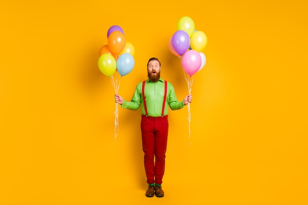 Foto in voller Länge von erstaunten imposanten charmanten Mann Hipster gehen Party halten viele Ballons beeindruckt festlichen Ereignis schreien wow omg tragen stilvolle Kleidung über gelbe Farbe isoliert