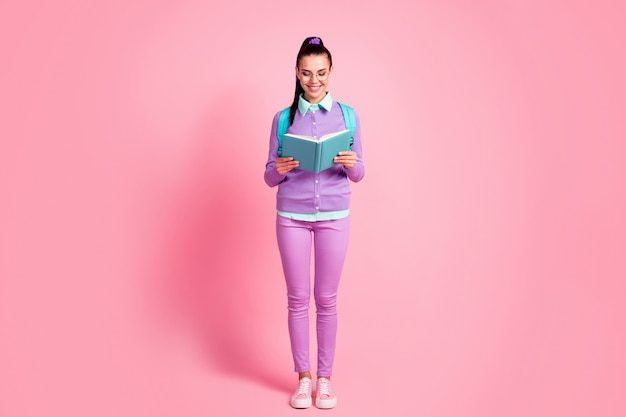 Foto in voller Länge von junger Dame halten Look-Buch strahlendes Lächeln tragen Specs Tasche violette Pullover Hose Turnschuhe isoliert rosa Hintergrund