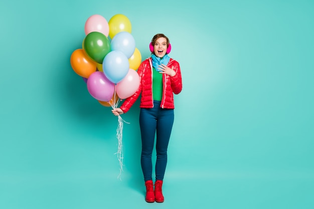 Foto in voller Länge von lustigen Emotionen Dame unerwartete Geburtstagsfeier viele bunte Luftballons überraschen tragen lässig roten Mantel Schal rosa Ohrenschützer Hosen Stiefel isoliert blaugrün Farbe Wand