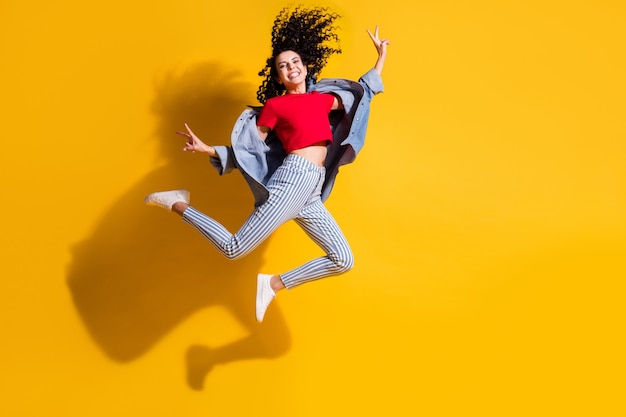 Foto in voller Länge von Mädchen Jump Show V-Zeichen tragen gestreifte Jeans abgeschnittene rote T-Shirt Jacke Turnschuhe isoliert gelber Hintergrund