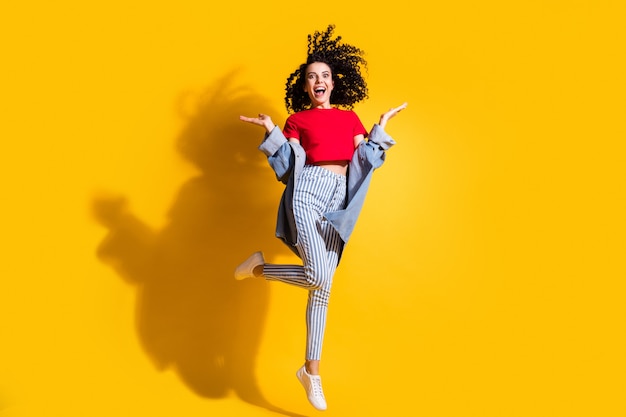 Foto in voller Länge von Mädchen springen Handflächen öffnen Mund tragen gestreifte Jeans abgeschnittene rote T-Shirt Jacke Turnschuhe isoliert gelber Hintergrund