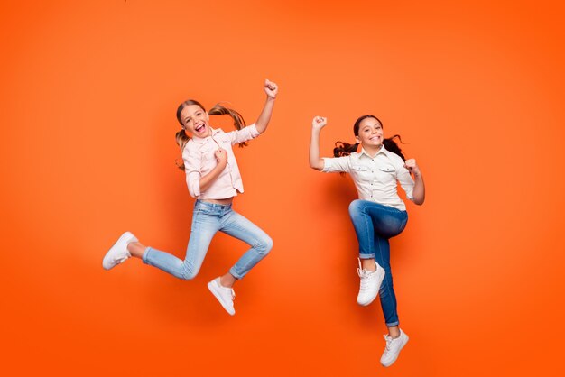 Foto in voller Länge von positiv freudig zwei Freundinnen springen Ruhe entspannen gewinnen Schule Sportwettbewerb erhöhen Fäuste tragen weißes Hemd Denim Jeans über orange Farbe Hintergrund isoliert