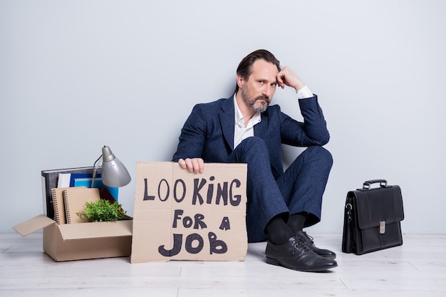 Foto in voller Länge von Verlierer-Arbeiter reifer alter Mann arbeitsloser Mann halten Plakat brauchen Arbeit sitzen Boden Aktentasche Sachen Box Sachen verloren alles Anzug Schuhe isoliert grauen Hintergrund tragen