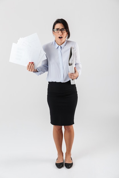 Foto in voller Länge von verwirrter Geschäftsfrau, die Brillen hält, die Papierordner im Büro halten, lokalisiert über weißer Wand