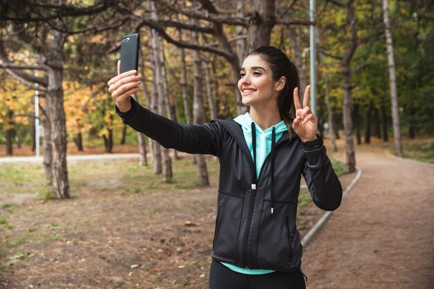 Foto de increíble joven mujer bonita fitness al aire libre en el parque con teléfono móvil tomar un selfie con gesto de paz.