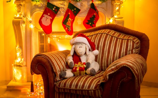 Foto interior de cordero de juguete sentado en una silla junto a la chimenea en Navidad