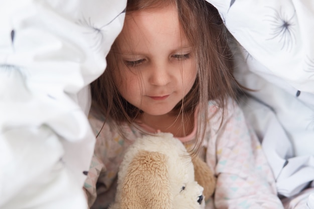 Foto interior da adorável criança menina brincando com ursinho de pelúcia na cama sob o cobertor