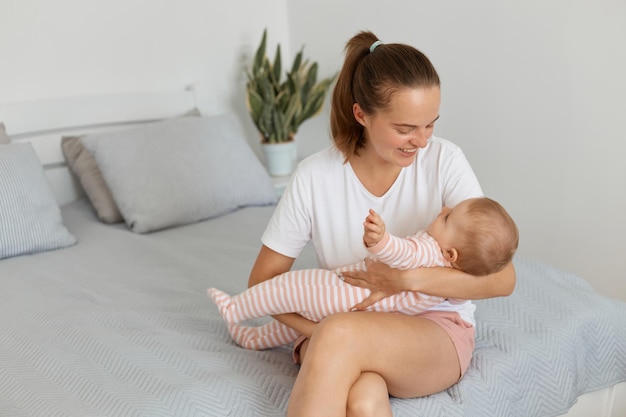 Foto interior da jovem mãe feliz vestindo camiseta branca sentada na cama com a filha brincando com criança criança vestindo dorminhoco listrado expressando felicidade