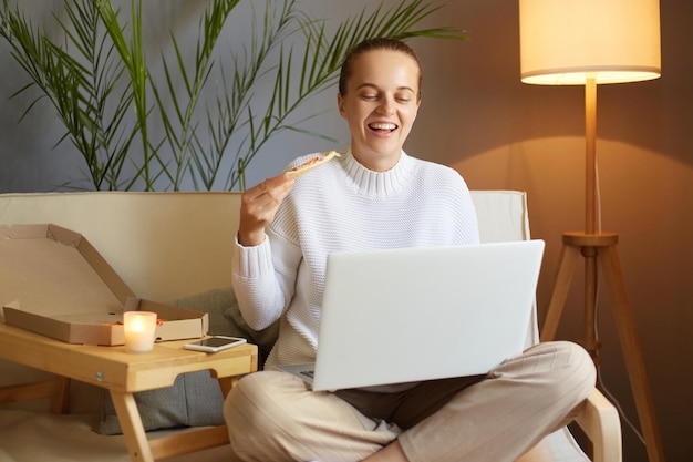 Foto interior de alegre e otimista linda mulher vestindo jumper branco sentado na tosse e trabalhando no laptop ou assistindo filme comendo fast food desfrutando de pizza