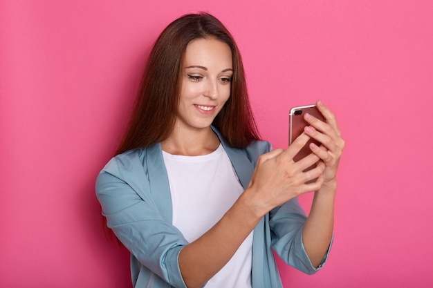 Foto interior de encantada doce adorável jovem segurando o smartphone em ambas as mãos, fazendo selfie