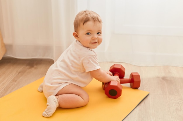 Foto interior de encantadora criança bonita sentada no tapete de ioga com halteres olhando para a câmera com sorriso explorando equipamentos esportivos